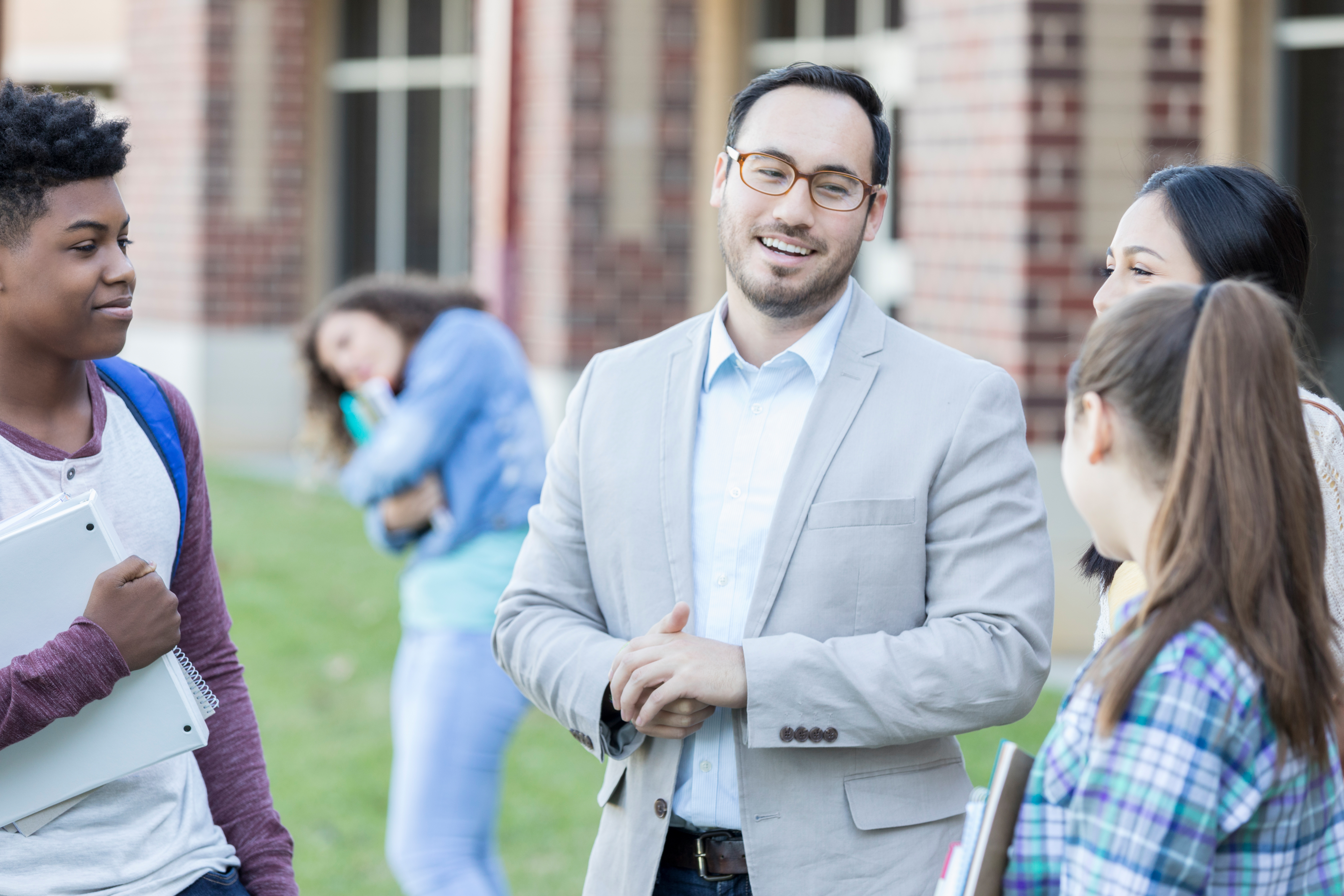 Engaged male teacher talking with diverse high school students outside a school building, representing the power of educator connection and student engagement.